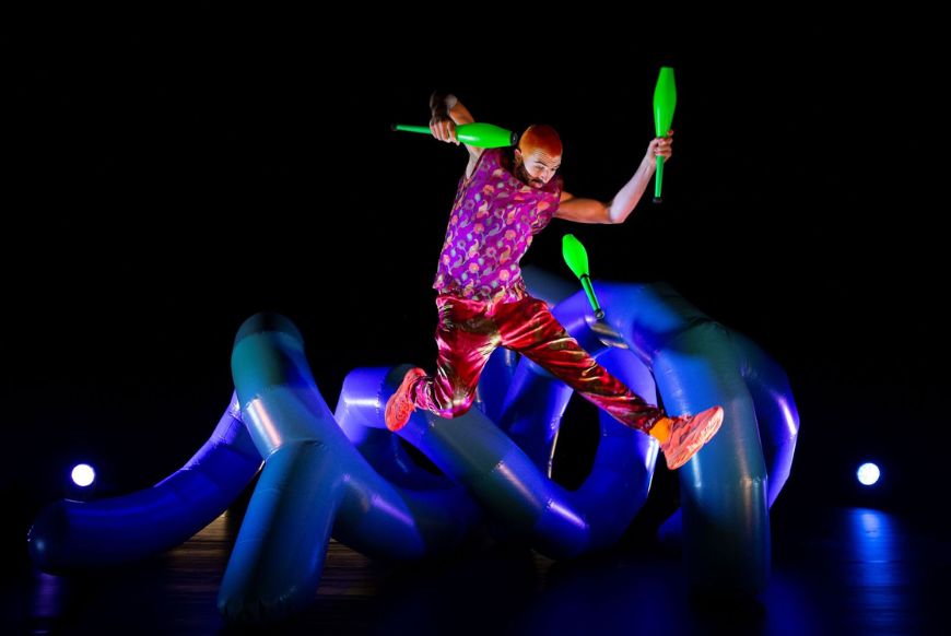 a man jumping while juggling neon green bwoling pins, with lots of inflatable blue tubes behind him